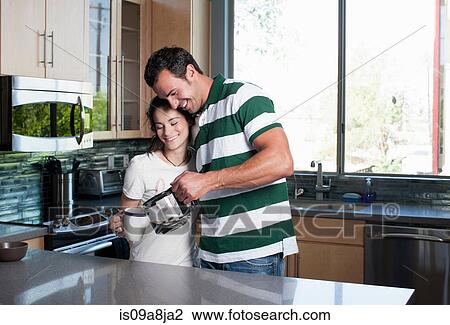 Stock Image - Young couple pouring coffee from pot in kitchen, smiling. Fotosearch