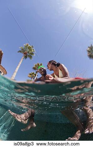 Stock Image - Young couple sitting at the edge of swimming pool, surface level view. Fotosearch