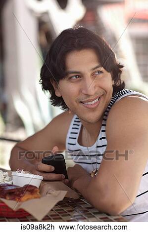 Stock Image - Young man holding mobile phone in outdoor cafe, looking away. Fotosearch