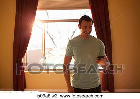 Young man looking at mobile phone in hotel room, smiling View Large Photo Image Stock Image - Young man looking at mobile phone in hotel room, smiling. Fotosearch