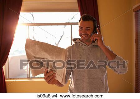 Picture - Young man looking at newspaper and using mobile phone in hotel room, smiling. Fotosearch