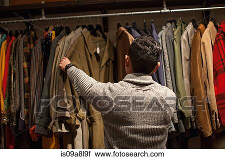 Stock Image - Young man looking through clothes rail in vintage shop. Fotosearch
