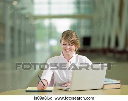 Stock Image - Young student studying taking notes and using laptop, smiling. Fotosearch