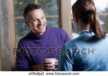Stock Image - Young woman and mature man holding hot drink indoors, smiling. Fotosearch