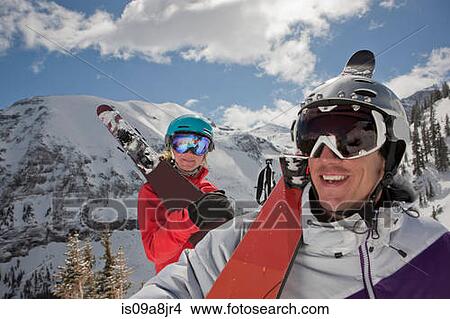 Picture - Young woman and mid adult man in skiwear carrying skis on shoulder, smiling. Fotosearch