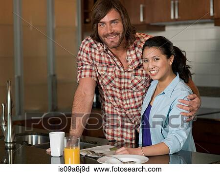 Stock Image - Young woman and mid adult man smiling in kitchen, portrait. Fotosearch