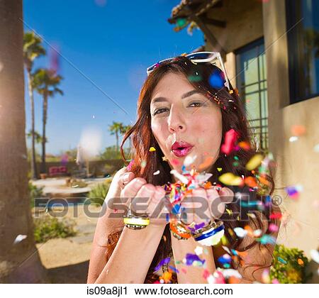 Stock Image - Young woman blowing confetti, portrait. Fotosearch