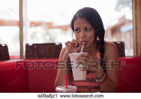 Stock Image - Young woman drinking milkshake in diner, looking away. Fotosearch