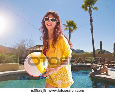 Stock Photo - Young woman holding beach ball at poolside, portrait. Fotosearch