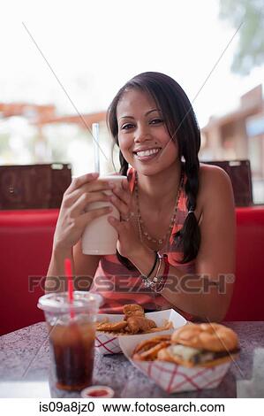 Stock Image - Young woman holding milkshake in diner, smiling. Fotosearch