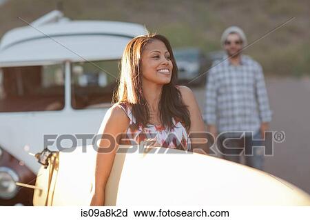 Stock Image - Young woman holding surfboard on road trip, smiling. Fotosearch