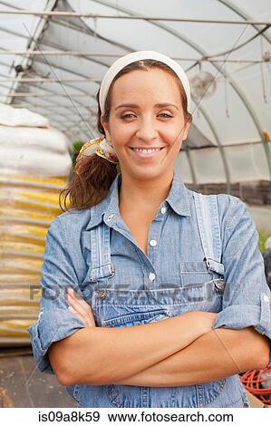 Stock Photo - Young woman in dungarees smiling in garden centre, portrait. Fotosearch