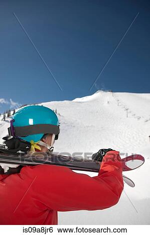 Stock Photograph - Young woman in skiwear holding skis over shoulder at the bottom of ski slope. Fotosearch