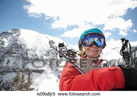 Stock Photography - Young woman in skiwear holding skis over shoulder, smiling. Fotosearch