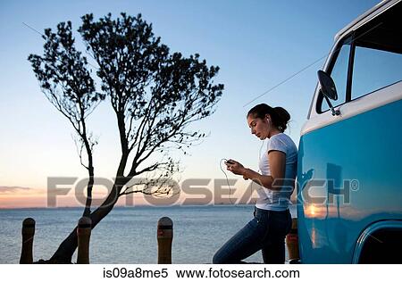 Stock Photography - Young woman leaning on camper van at dusk. Fotosearch