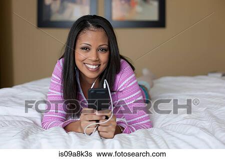 Stock Photograph - Young woman lying on bed and listening to mp3 player, portrait. Fotosearch