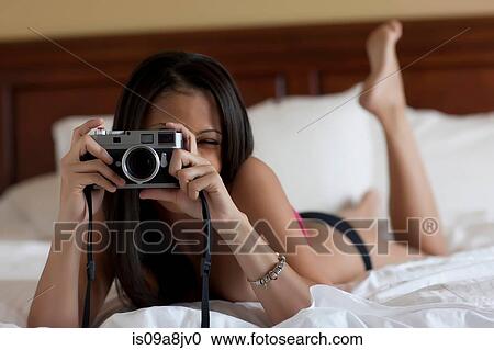 Stock Image - Young woman lying on bed in underwear taking photograph. Fotosearch