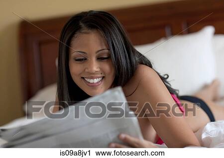 Stock Image - Young woman lying on bed reading newspaper. Fotosearch