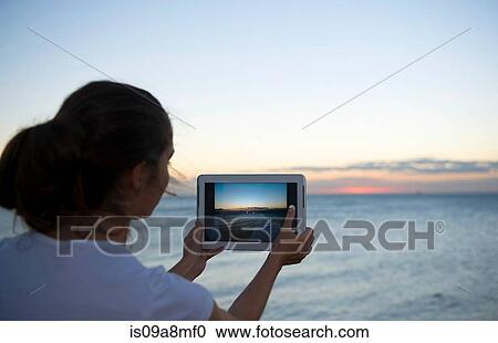Stock Image - Young woman photographing with her digital tablet at sunset. Fotosearch