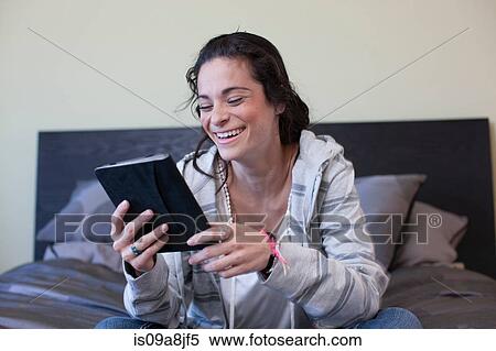 Stock Photography - Young woman sitting bed reading electronic book, smiling. Fotosearch