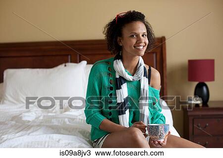 Stock Image - Young woman sitting on bed holding cup, portrait. Fotosearch