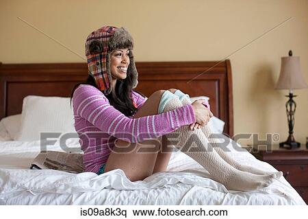 Stock Photography - Young woman sitting on bed wearing ushanka, smiling. Fotosearch