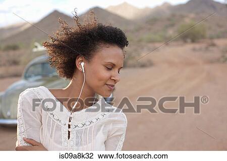 Stock Image - Young woman wearing earphones in desert on road trip, smiling. Fotosearch