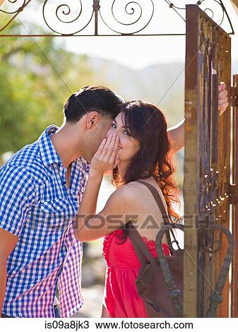 Stock Image - Young woman whispering to man by iron gate, portrait. Fotosearch