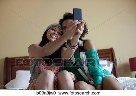 Stock Image - Young women sitting on bed taking self portrait with mobile phone. Fotosearch