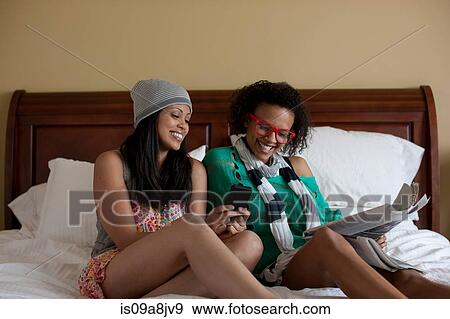 Young women sitting on bed using mobile phone, laughing View Large Photo Image Stock Photo - Young women sitting on bed using mobile phone, laughing. Fotosearch