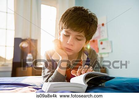 Stock Image - Boy lying on bed reading book. Fotosearch
