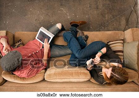 Stock Photography - Couple sitting on sofa using digital tablet and smartphone. Fotosearch