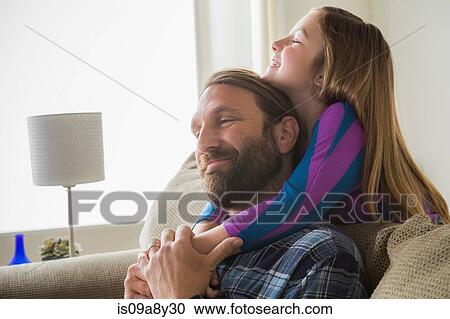 Stock Image - Daughter hugging father from behind sofa. Fotosearch