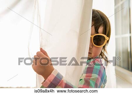 Stock Image - Girl peering round curtain wearing sunglasses. Fotosearch