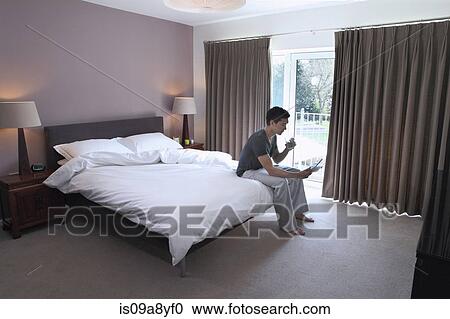 Man sitting on bed looking at digital tablet View Large Photo Image Stock Image - Man sitting on bed looking at digital tablet. Fotosearch