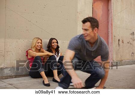 Stock Image - Man skating past women sitting on ground. Fotosearch
