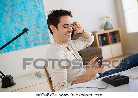 Stock Image - Mid adult man on cell phone in home office. Fotosearch