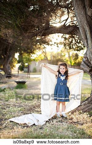Stock Photo - Portrait of girl holding sheet with arms out under tree. Fotosearch