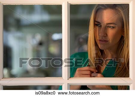 Stock Image - Portrait of young woman looking through window. Fotosearch