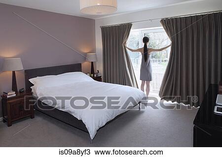 Stock Image - Woman drawing open curtains of bedroom. Fotosearch