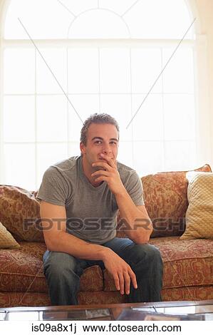 Stock Image - Young man sitting on sofa with hand on chin. Fotosearch
