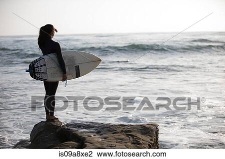 Stock Image - Young woman standing on rocks with surfboard. Fotosearch