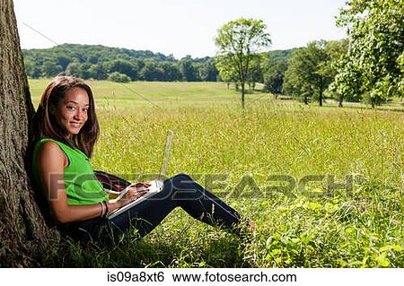 Stock Photograph - Young woman using laptop sitting by tree. Fotosearch