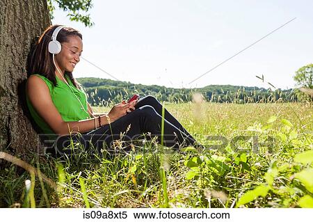 Stock Photography - Young woman wearing headphones sitting by tree. Fotosearch
