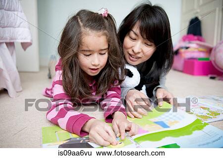 Stock Photograph - Girl lying on front and reading sticker book with mother. Fotosearch