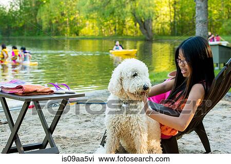 Stock Image - Girl sitting on chair by lake with dog. Fotosearch