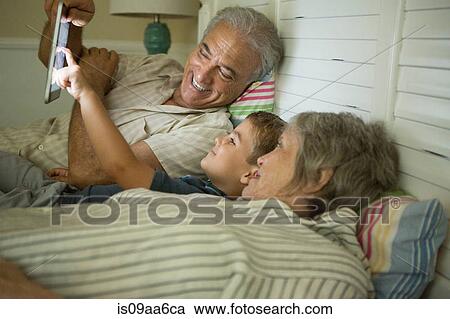 Stock Image - Grandparents lying down with grandson and looking at digital tablet. Fotosearch