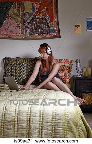 Stock Image - Mid adult woman sitting on bed using laptop. Fotosearch
