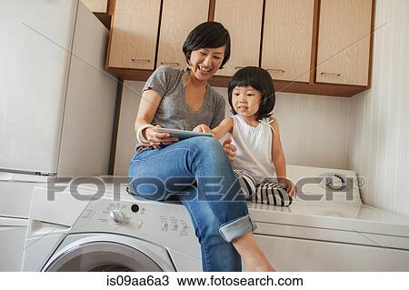 Stock Image - Mother and daughter sitting on washing machine using tablet. Fotosearch
