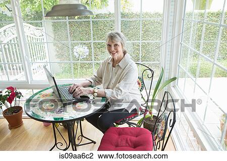Stock Image - Senior woman using latpop in conservatory, portrait. Fotosearch